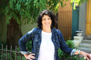 Picture of Jane Pfieffer, white woman with dark curly hair in a jean jacket, standing outside near a fence.